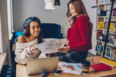 Happy female fashion designers discussing ideas for clothing sketches near desktop at bookstore, smiling african hipster girl sitting at table with laptop and talking about illustration with friend